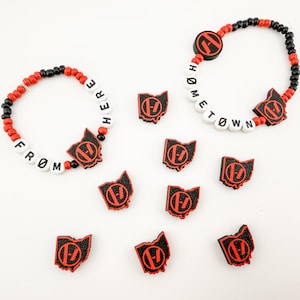 May include: Two beaded bracelets featuring alternating red and black beads, white letter beads spelling FROM HERE and HOMETOWN, and black and red Ohio-shaped charms. Seven additional black and red Ohio-shaped charms are arranged below the bracelets.
