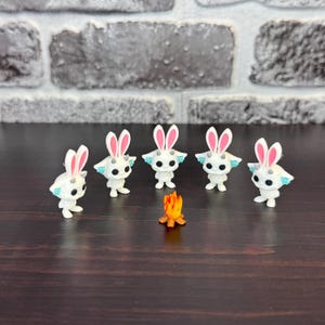 May include: Five white figurines with bunny ears and blue accents surround a small orange campfire. The figurines have black eyes and are posed as if they are gathered around the fire. The background is a brick wall and a dark wooden surface.