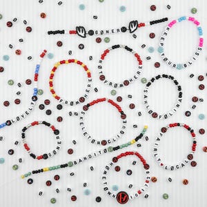 May include: A collection of handmade beaded bracelets and necklaces featuring white letter beads spelling phrases like GØNER, KEEP YOUR BLISS, FROM HERE, SKELETON CLIQUE, SAHLØ FØLINA, LOVELY, BANDITØ, FAIRLY LØCAL, and HØMETØWN, accented with red, black, and colorful beads.