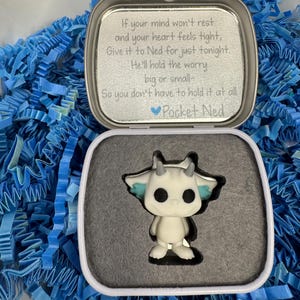 May include: A white, stylized figurine of a creature with horns and blue accents inside an open, silver tin. The tin contains a poem about worry and is surrounded by blue paper shreds. The text on the tin reads "Pocket Ned".
