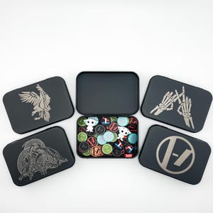 May include: A collection of black metal tins, some open, filled with colorful tokens and small figurines. The tins feature various designs, including an eagle, a dragon, skeleton hands, and a band logo. The tokens have various colors and symbols.