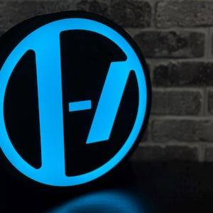 May include: A black, circular sign with a glowing blue logo. The logo features a stylized 'T' and a horizontal line, set against a black background. The sign is likely a decorative item or merchandise.