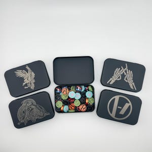 May include: A collection of black metal tins, some open and some closed, filled with colorful round objects. The closed tins feature various designs, including an eagle, a snake, and skeletal hands. The open tin reveals a variety of small, circular items.