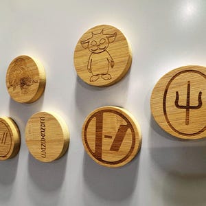 May include: A collection of round, wooden magnets with engraved designs. The magnets feature various images and symbols, including a cartoon character, a trident, and band logos. The magnets are attached to a white surface.