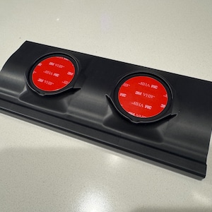 May include: A black plastic component with two circular recessed areas. Each area contains a red, circular adhesive pad with white text. The component has a rectangular shape with curved edges and a slightly raised lip along the bottom.