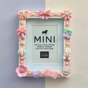 May include: A rectangular photo frame with a white interior and a pink border adorned with decorative elements. The frame features a butterfly, flowers, a bow, a hot air balloon, and the word "Sweet". The frame is 3 inches x 4 inches / 7.6 cm x 10.1 cm.