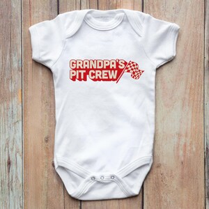 Peut inclure: Combinaison blanche pour bébé avec le texte rouge "Grandpa's Pit Crew" et un drapeau à damier rouge et blanc.