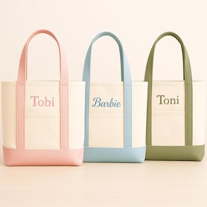May include: Three canvas tote bags with cream-colored bodies, each featuring a different colored base, handles, and trim. The bags are personalized with the names "Tobi," "Barbie," and "Toni" in coordinating colors.