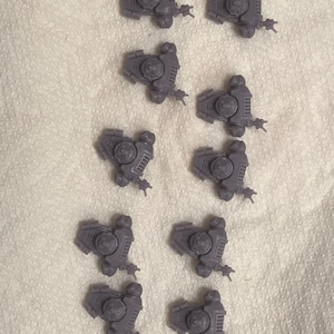 May include: Ten gray plastic miniature vehicle models are arranged on a white, textured surface. Each model has a detailed design with a central turret, side panels, and small weapon attachments. The models are likely for tabletop gaming or model building.