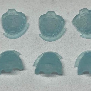 May include: Ten translucent, light blue plastic components are arranged on a white surface. The components have a rounded top with a flat base and a curved, wing-like extension. The objects are evenly spaced and appear to be identical.