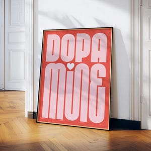 May include: A framed art print with the word "Dopamine" in large, light pink letters on a coral background. A small white heart is between the words. The print is in a black frame and leans against a white wall and wooden floor.