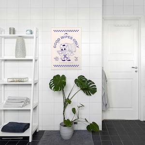 May include: A framed poster with a cartoon toilet paper roll character in purple and white. The poster reads "Good Wipes Only" and is displayed in a bathroom setting with a white shelf, a potted plant, and a white door.
