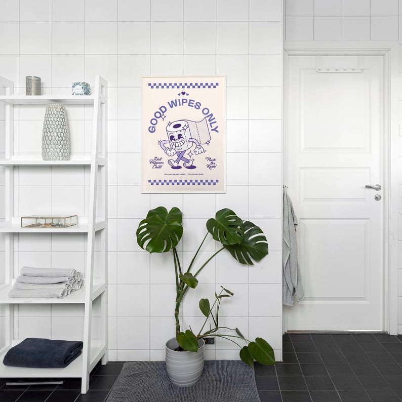 May include: A framed poster with a cartoon toilet paper roll character in purple and white. The poster reads "Good Wipes Only" and is displayed in a bathroom setting with a white shelf, a potted plant, and a white door.