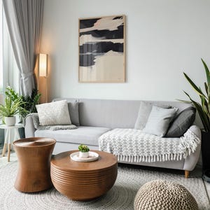 May include: A contemporary living room featuring a grey sofa with cushions and a patterned throw. Two wooden coffee tables and a knitted pouffe rest on a grey rug. An abstract artwork hangs above the sofa, with a floor lamp providing ambient light.