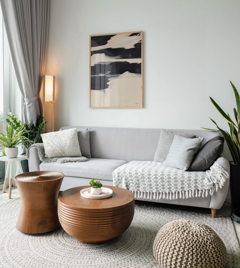 May include: A contemporary living room featuring a grey sofa with cushions and a patterned throw. Two wooden coffee tables and a knitted pouffe rest on a grey rug. An abstract artwork hangs above the sofa, with a floor lamp providing ambient light.