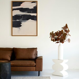 May include: A contemporary living room with a brown leather sofa, a black side table, and a white, sculptural side table. An abstract painting with black, white, and pink brushstrokes hangs on the wall. A white vase holds reddish-brown foliage.