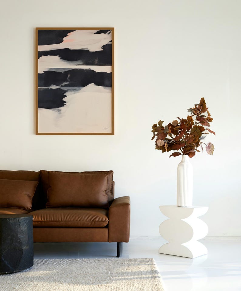 May include: A contemporary living room with a brown leather sofa, a black side table, and a white, sculptural side table. An abstract painting with black, white, and pink brushstrokes hangs on the wall. A white vase holds reddish-brown foliage.