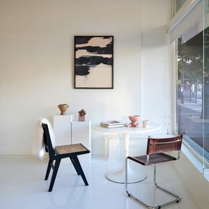 May include: A contemporary interior with a black and white abstract artwork, a black chair with a woven seat, and a brown leather chair. A small white table displays decorative items. The room has white walls and a large window.