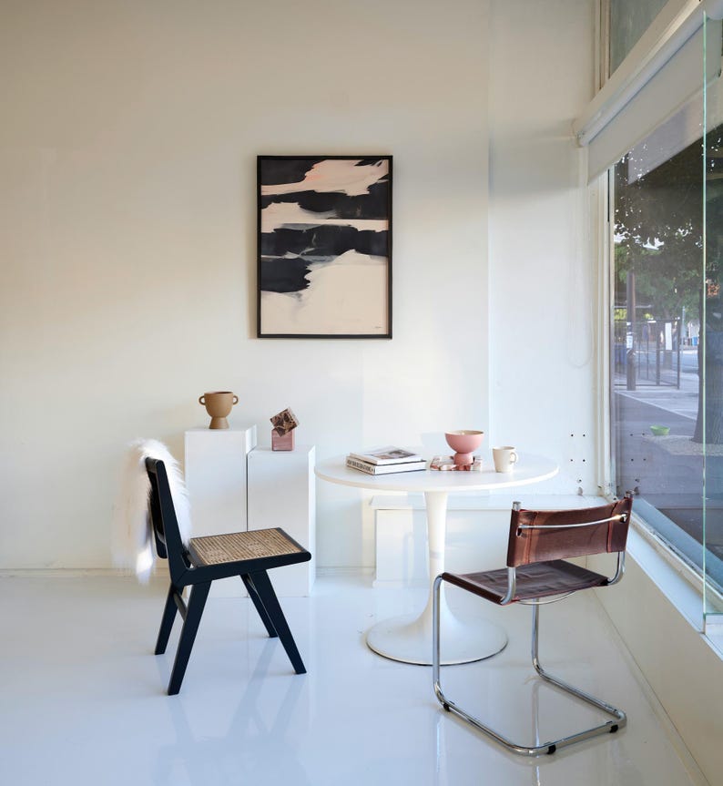 May include: A contemporary interior with a black and white abstract artwork, a black chair with a woven seat, and a brown leather chair. A small white table displays decorative items. The room has white walls and a large window.