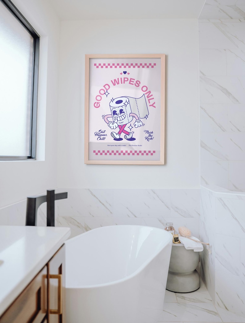 May include: A framed print with a cartoon toilet paper roll character that reads "Good Wipes Only." The print has a light wood frame and is displayed in a bathroom with a white bathtub and marble tile.