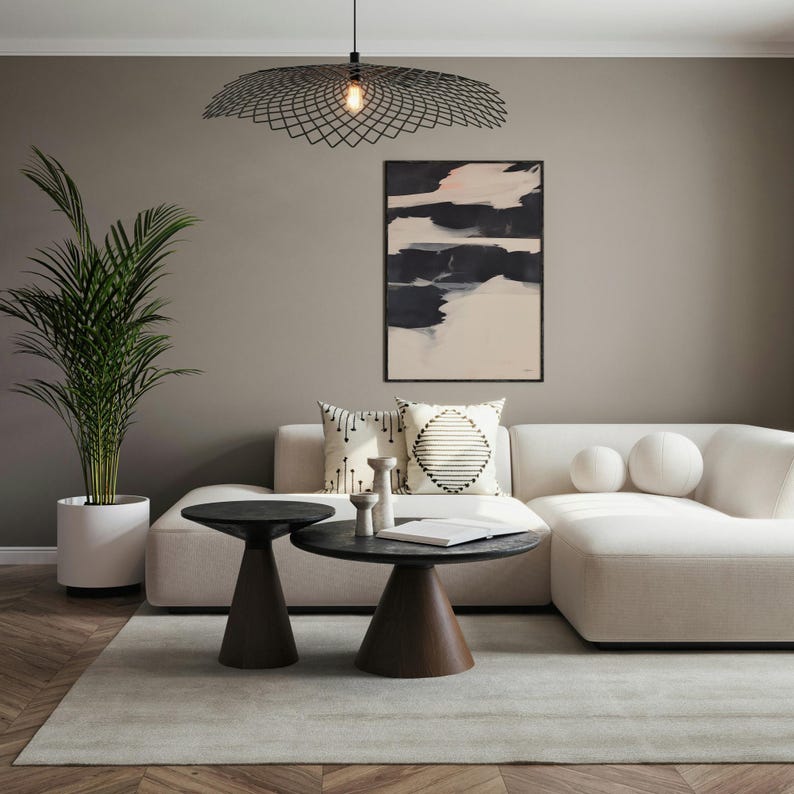 May include: A contemporary living room featuring a cream-coloured sofa, two dark brown side tables, and an abstract painting. A black geometric pendant light hangs overhead. A potted palm and decorative cushions complete the scene.