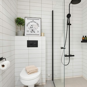 May include: A modern bathroom with white tiled walls and a glass shower enclosure. A framed black and white print that says "GOOD WIPES ONLY" hangs above the toilet. A roll of toilet paper and a folded towel are also visible.