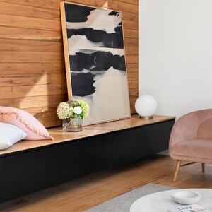 May include: An abstract painting with black and white brushstrokes in a gold frame. The artwork is displayed above a black cabinet with a wooden top. A pink cushion, a vase of flowers, and a white globe lamp are also visible.