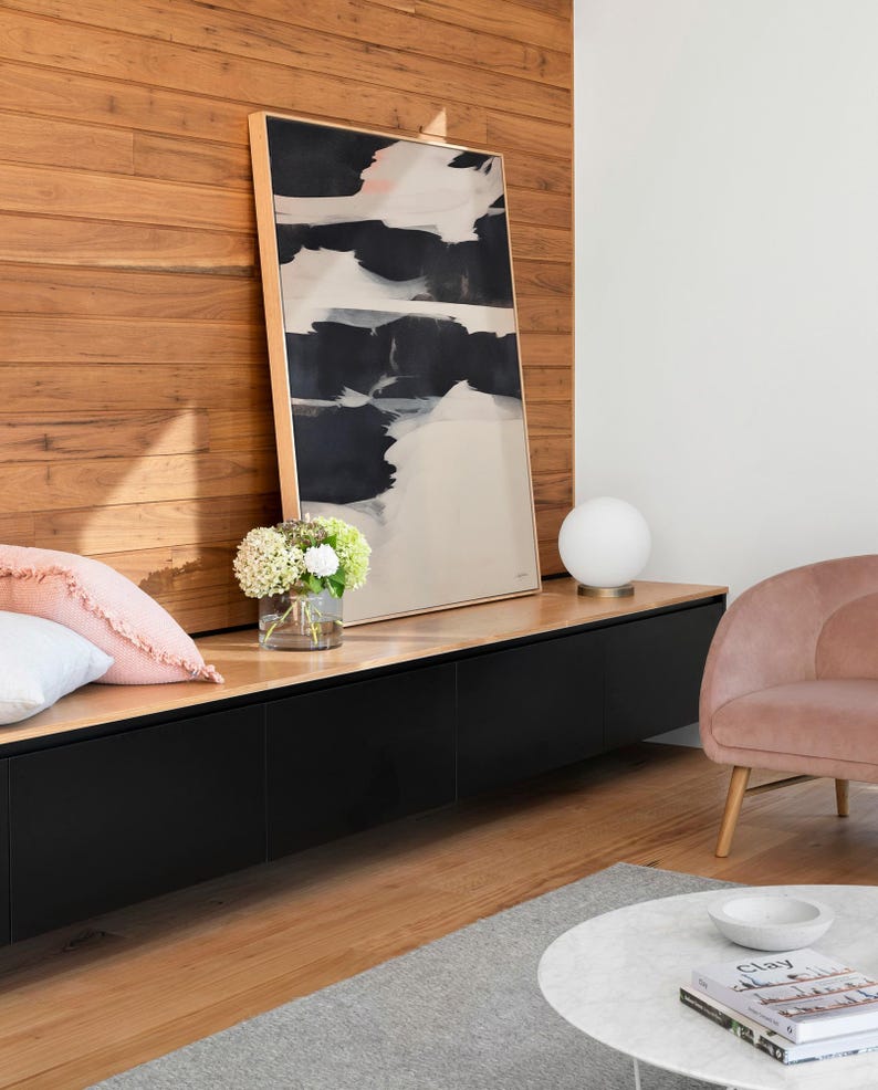 May include: An abstract painting with black and white brushstrokes in a gold frame. The artwork is displayed above a black cabinet with a wooden top. A pink cushion, a vase of flowers, and a white globe lamp are also visible.