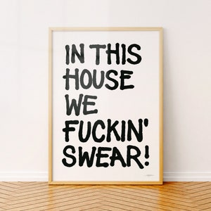 Op de afbeelding: Een ingelijste print met de tekst "IN THIS HOUSE WE FUCKIN' SWEAR!" in vette zwarte letters op een witte achtergrond. De lijst heeft een lichte houtkleur en de print wordt op een houten vloer weergegeven.
