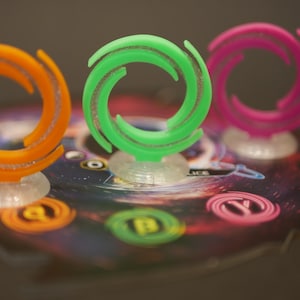 Op de afbeelding: Drie kleurrijke plastic spelstukken in de vorm van een draaiende vortex. De stukken zijn oranje, groen en roze en staan op een spelbord met een zwart-witte galaxy-ontwerp.