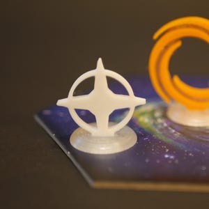 Puede incluir: Una pieza de juego blanca en forma de estrella con un marco circular, colocada sobre una pequeña base transparente. La pieza está colocada sobre un tablero de juego con un diseño temático espacial, con una galaxia de azules, morados y estrellas blancas. Una pieza de juego naranja en forma de anillo está en el fondo.