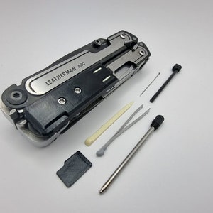 May include: A multi-tool with the text "LEATHERMAN ARC" on a silver plate. The tool is black and silver, with several small tools and accessories laid out beside it, including a small screwdriver and tweezers.