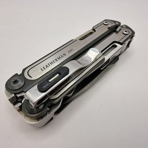 Op de afbeelding: Een zilver en zwart multi-tool met de tekst "LEATHERMAN ARC". De tool heeft een zakclip. De afbeelding is een close-up, die het ontwerp en de componenten van de tool benadrukt. De tool is waarschijnlijk van metaal.