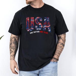 May include: A black short-sleeved t-shirt featuring the text USA in large block letters filled with red, white, and blue patriotic graphics including stars, stripes, and landmarks. Below the text, the words ONE NATION ONE TEAM are printed.