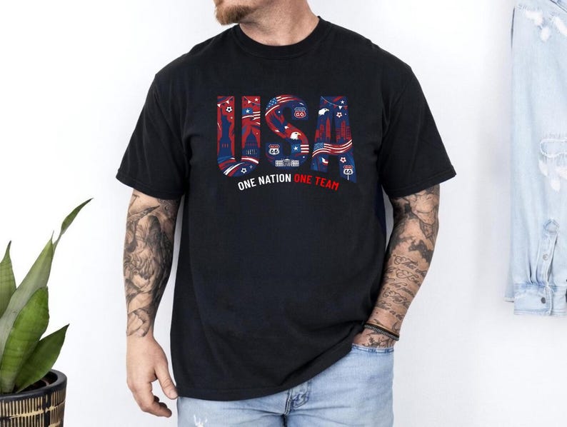 May include: A black short-sleeved t-shirt featuring the text USA in large block letters filled with red, white, and blue patriotic graphics including stars, stripes, and landmarks. Below the text, the words ONE NATION ONE TEAM are printed.