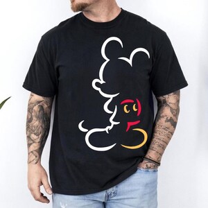 May include: Black t-shirt featuring a white outline of Mickey Mouse's face and body. The design includes red, yellow, and white accents. The shirt is short-sleeved and appears to be made of a soft material.