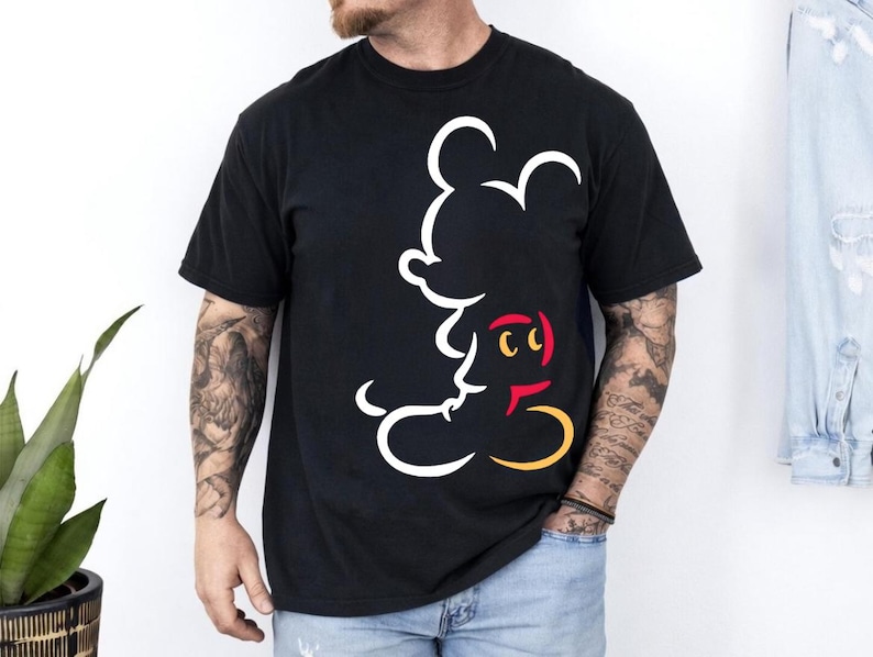 May include: Black t-shirt featuring a white outline of Mickey Mouse's face and body. The design includes red, yellow, and white accents. The shirt is short-sleeved and appears to be made of a soft material.