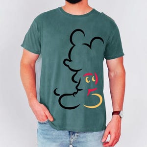 May include: Teal-colored t-shirt featuring a black outline of a cartoon mouse. The mouse has red, yellow, and black accents. The shirt is a short-sleeved crew neck.