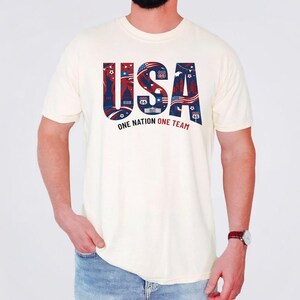 May include: A cream-colored short-sleeve t-shirt featuring the text USA in large blue letters filled with red, white, and blue patriotic patterns, including stars, stripes, and landmarks. Below the text is the phrase ONE NATION ONE TEAM.