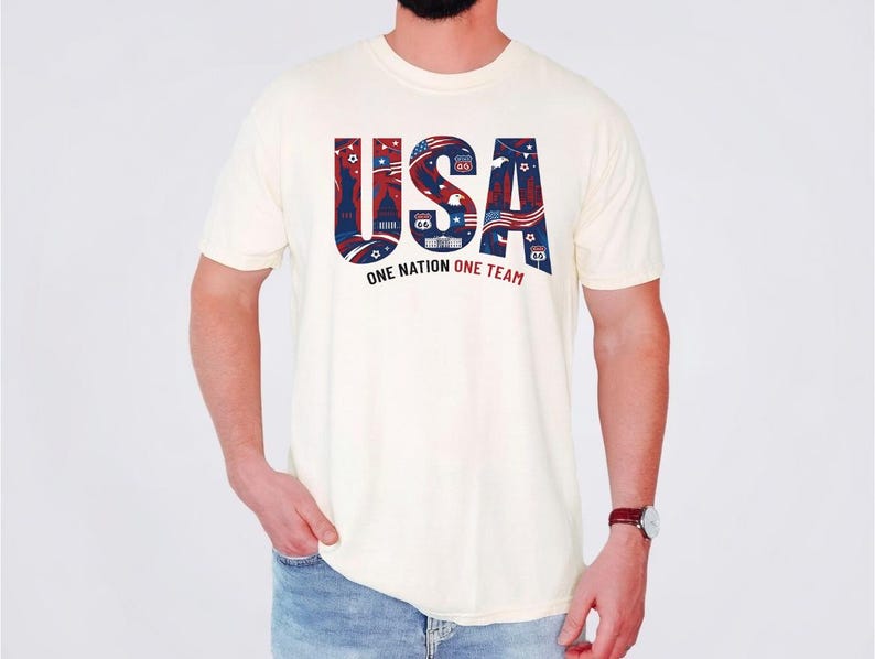 May include: A cream-colored short-sleeve t-shirt featuring the text USA in large blue letters filled with red, white, and blue patriotic patterns, including stars, stripes, and landmarks. Below the text is the phrase ONE NATION ONE TEAM.