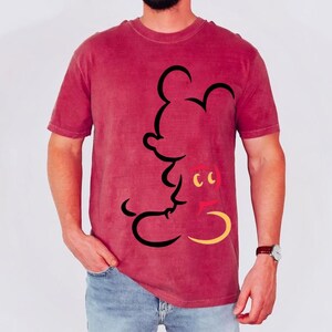 May include: A maroon t-shirt featuring a black outline of Mickey Mouse. The cartoon character's face is detailed with yellow and red accents. The shirt is made of a soft material and has short sleeves.
