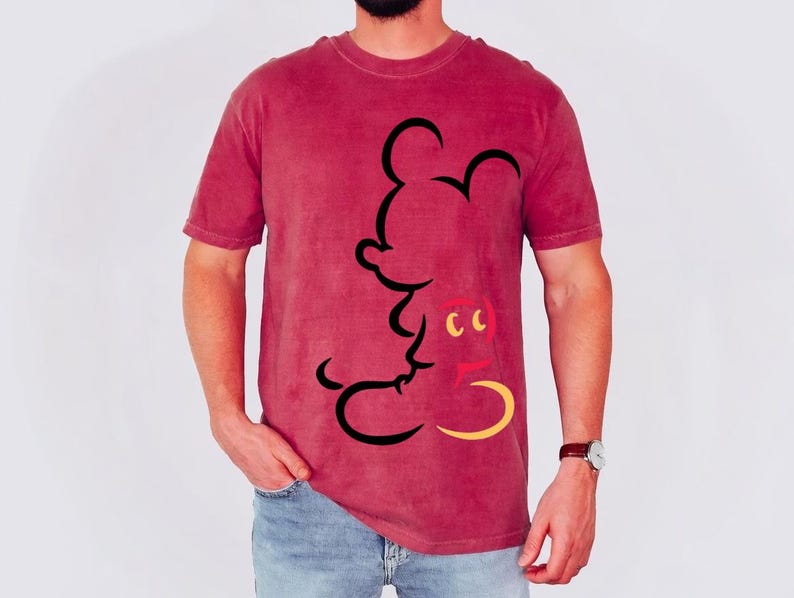 May include: A maroon t-shirt featuring a black outline of Mickey Mouse. The cartoon character's face is detailed with yellow and red accents. The shirt is made of a soft material and has short sleeves.