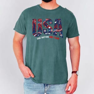 May include: A dark teal short-sleeve t-shirt featuring the text USA in large block letters filled with red, white, and blue patriotic graphics including stars, stripes, and landmarks. Below the text is the phrase ONE NATION ONE TEAM in white and red.