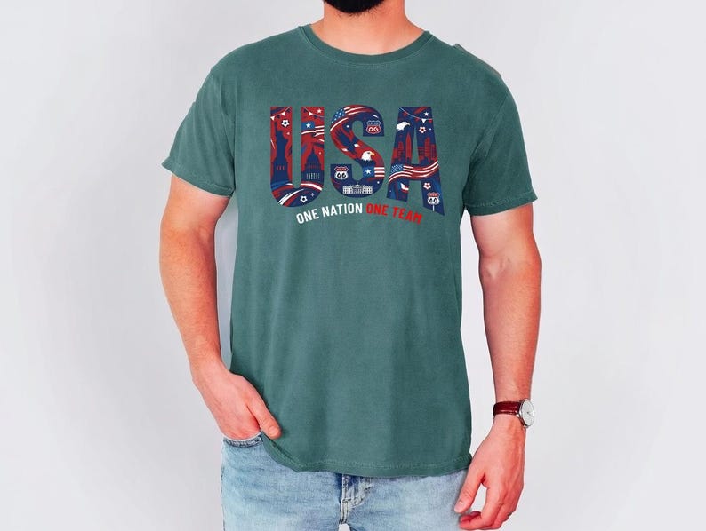 May include: A dark teal short-sleeve t-shirt featuring the text USA in large block letters filled with red, white, and blue patriotic graphics including stars, stripes, and landmarks. Below the text is the phrase ONE NATION ONE TEAM in white and red.