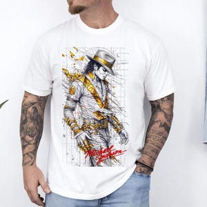 May include: A white short-sleeved t-shirt featuring a graphic illustration of Michael Jackson wearing a fedora and a gold-accented outfit. The design includes abstract gold lines, a grid background, and the name Michael Jackson in red script.