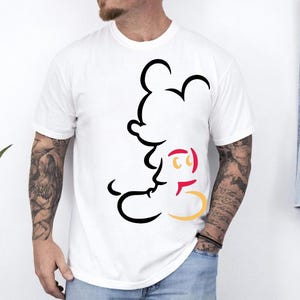 May include: White t-shirt featuring a black outline of Mickey Mouse. The design includes red and yellow accents. The shirt is a classic crew neck style.