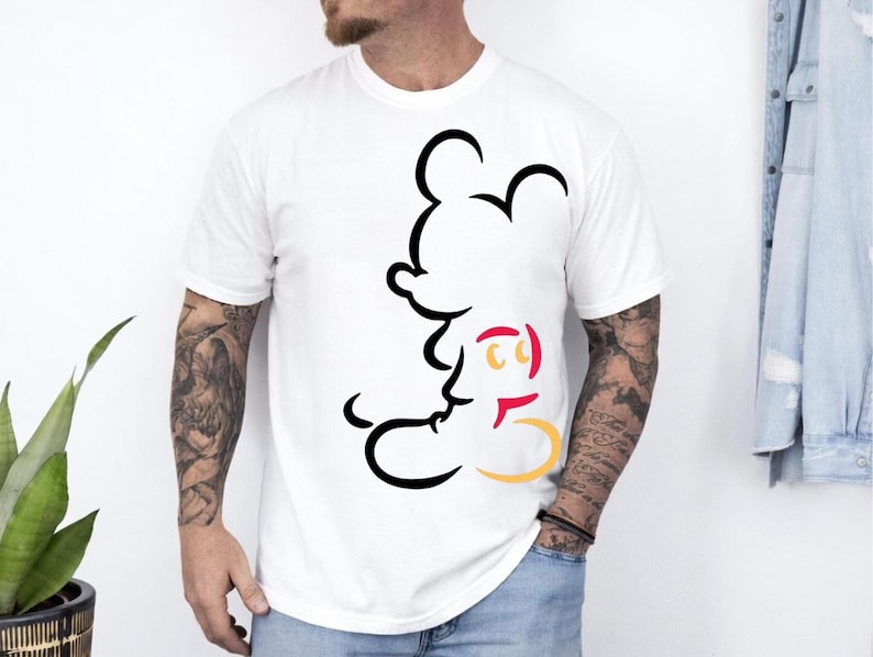 May include: White t-shirt featuring a black outline of Mickey Mouse. The design includes red and yellow accents. The shirt is a classic crew neck style.