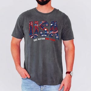 May include: A charcoal grey short-sleeve t-shirt featuring the large letters USA in a red, white, and blue graphic pattern containing patriotic symbols and Route 66 signs. Below the letters, the text reads ONE NATION ONE TEAM in white and red.