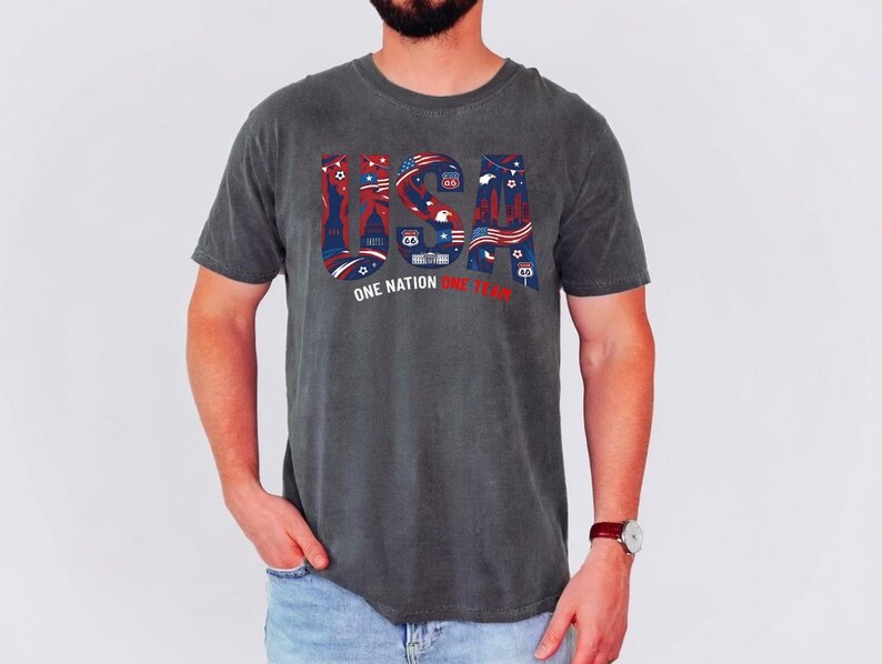 May include: A charcoal grey short-sleeve t-shirt featuring the large letters USA in a red, white, and blue graphic pattern containing patriotic symbols and Route 66 signs. Below the letters, the text reads ONE NATION ONE TEAM in white and red.
