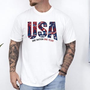 May include: A white short-sleeved t-shirt featuring the large blue and red text USA on the chest. The letters contain graphic illustrations of American landmarks, flags, and symbols. Below the text, the words ONE NATION ONE TEAM are printed.